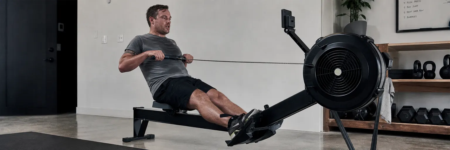 Adult mid-sprint on a rowing machine in a home gym, with kettlebells and dumbbells in the background